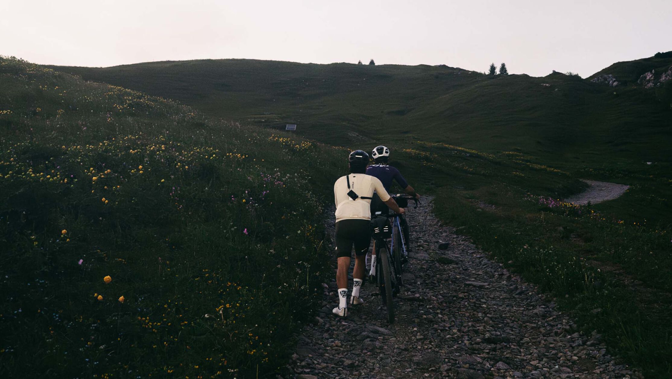 Gravel bikepacking setup&mdash;riders push their loaded bikes uphill through remote alpine trails, showcasing endurance and gear choices.